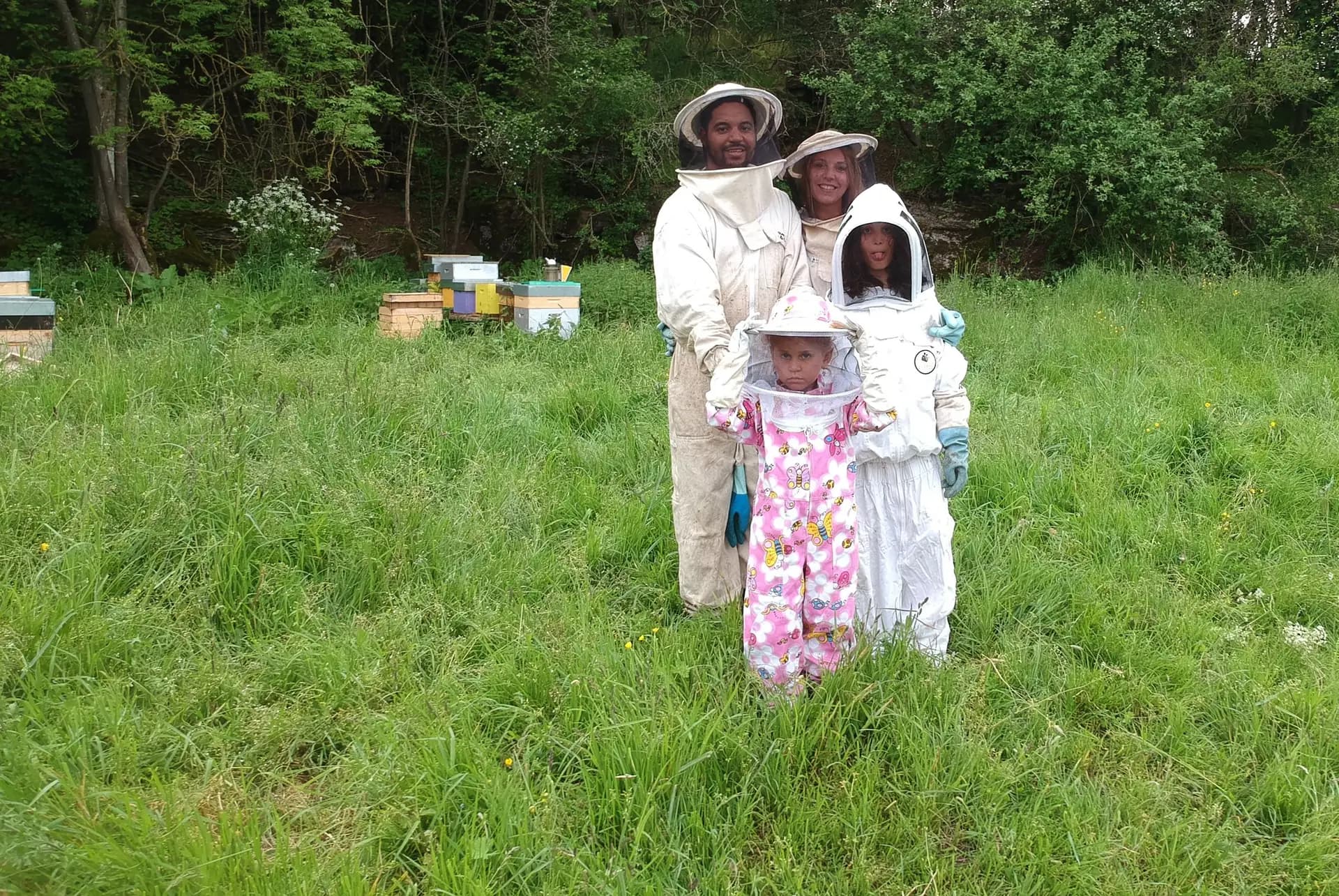 Une famille en tenue d’apiculteur, parents et enfants, dans une prairie devant des ruches.